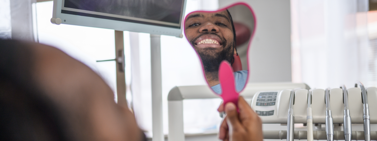Homem negro sorrindo ao se olhar em espelho de mão no dentista.