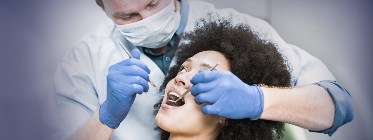  Dentista de máscara e luvas azuis examina a boca de uma paciente, que está deitada na cadeira de atendimento odontológico, enquanto segura um espelho dental e uma sonda exploradora.