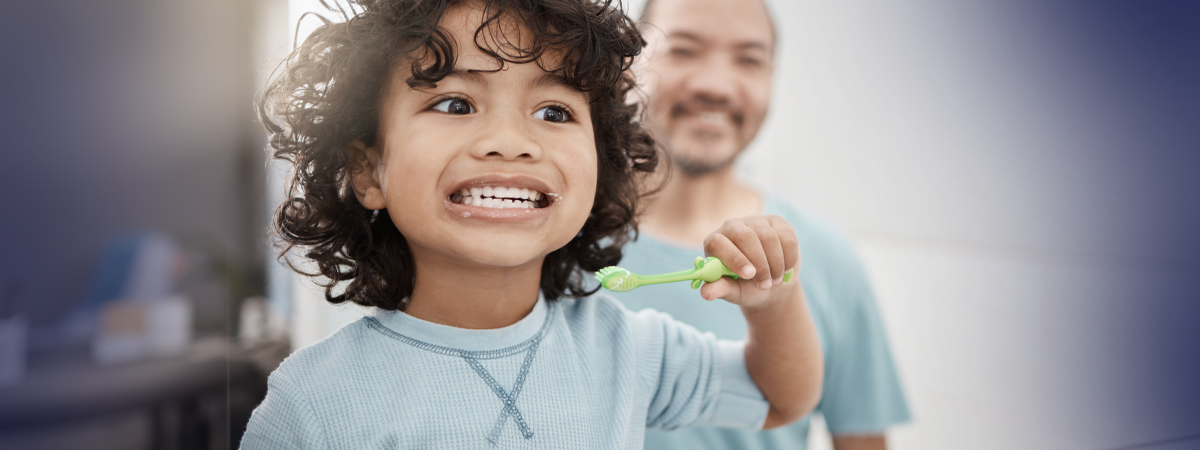Criança sorridente escovando os dentes com adulto ao fundo