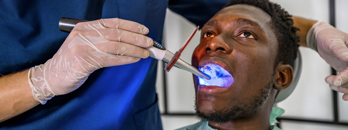 Homem negro recebendo tratamento dentário com luz azul em uma clínica.