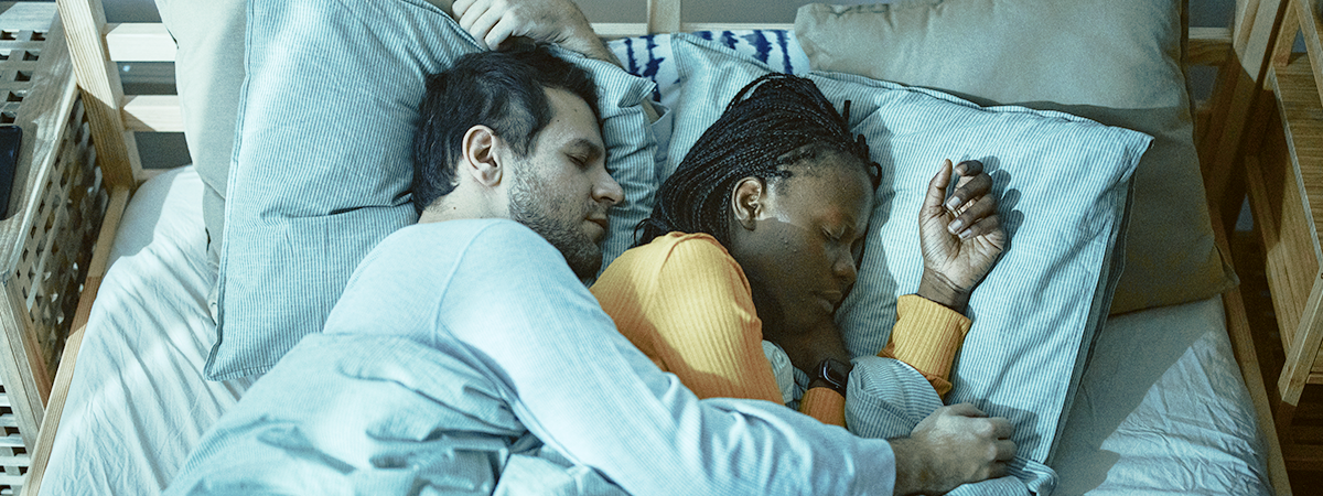 Casal dormindo abraçado na cama, homem branco com camisa azul e mulher negra com blusa laranja.
