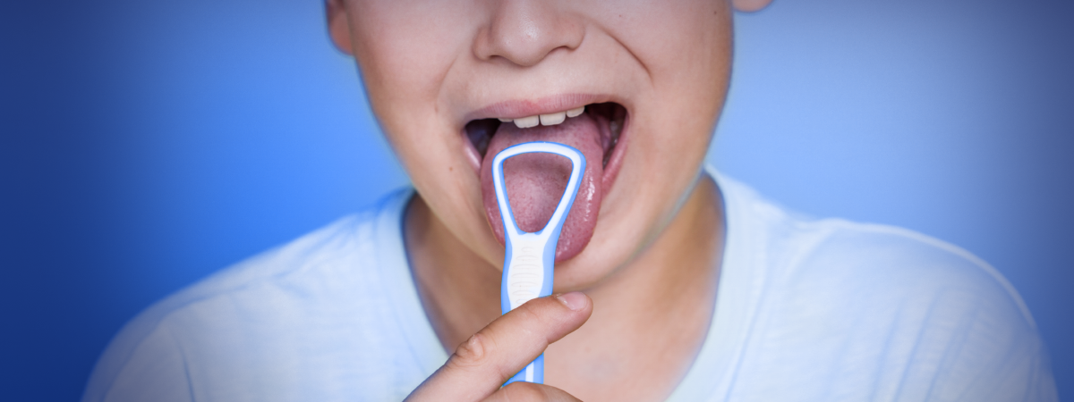 Uma criança sorridente mostra a língua através de um espelho de dentista, segurando um limpador de língua, em um fundo azul sólido.
