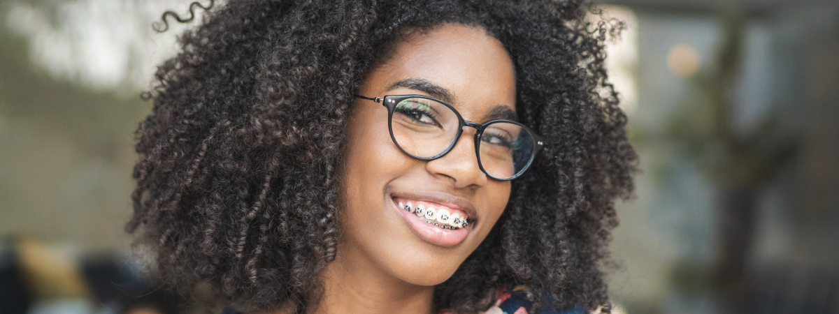 Mulher jovem sorridente com óculos e aparelho nos dentes, cabelo cacheado ao natural.