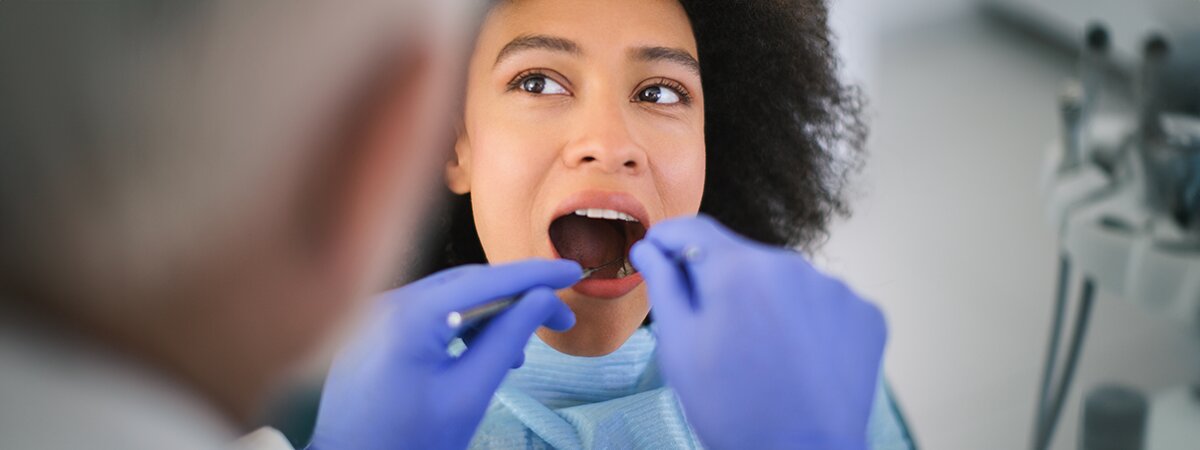 Mulher em consulta odontológica, abrindo a boca para o dentista com luvas azuis.