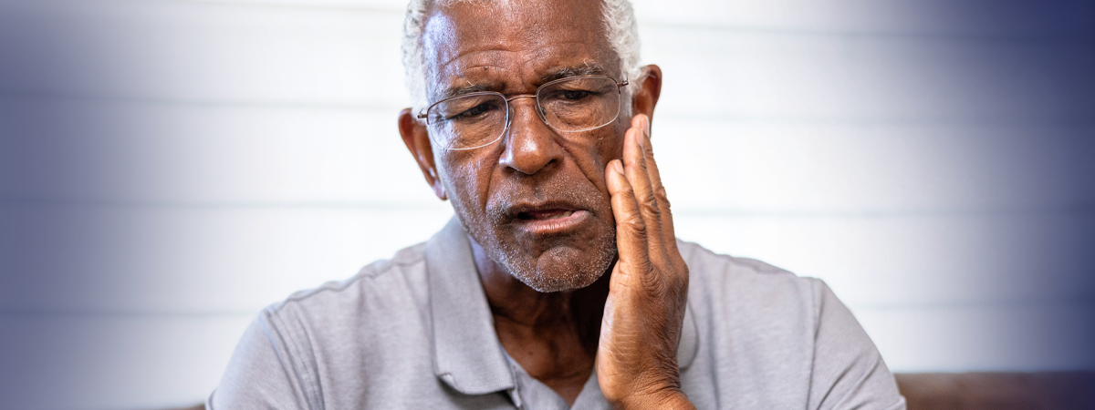 Homem mais velho com expressão de desconforto, mão no rosto, fundo listrado claro