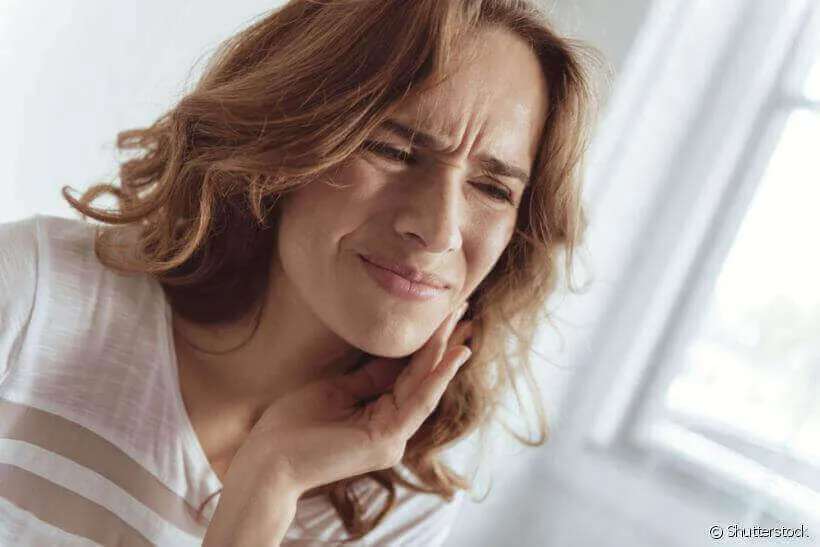 Mulher sentindo dor na boca, tocando a bochecha com uma expressão de desconforto, possivelmente devido a aftas.