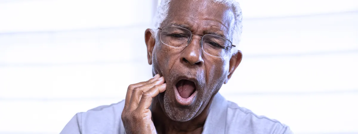 Um senhor idoso com óculos e camisa cinza aparenta bocejar ou sentir dor, segurando sua mandíbula com uma expressão de desconforto