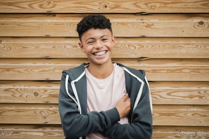 Adolescente sorridente com aparelho ortodôntico em frente a uma parede de madeira, representando os cuidados pós-tratamento ortodôntico