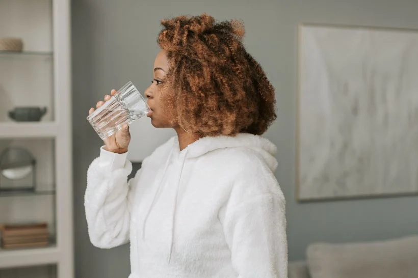 Mulher negra bebendo água de um copo transparente, vestindo um roupão branco felpudo. Ela está em pé, em um ambiente doméstico moderno, com decoração neutra e um quadro ao fundo, enfatizando a importância da hidratação para a saúde bucal pós-parto