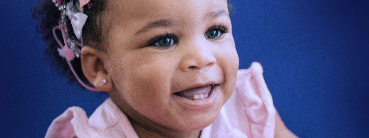 Bebê sorridente com pequenos dentinhos à mostra, usando uma faixa rosa na cabeça.