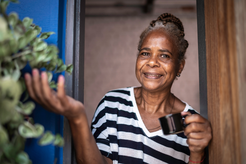 Senhora negra sorridente com xícara na mão ao lado de planta, porta aberta.