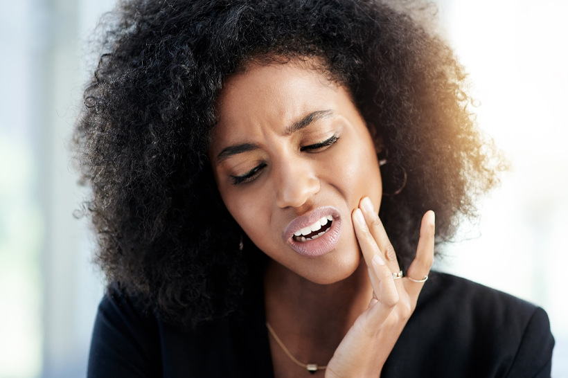Mulher negra jovem com dor de dente, mão no rosto, expressão de desconforto.