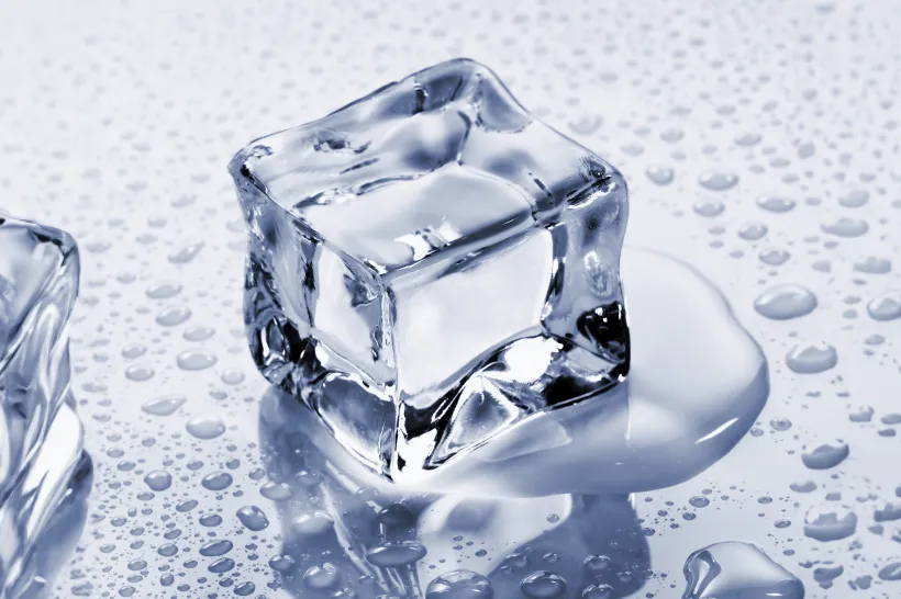 Cubos de gelo sobre uma superfície refletiva com gotas de água ao redor, sugerindo uma dica para aliviar dor de dente com frio.